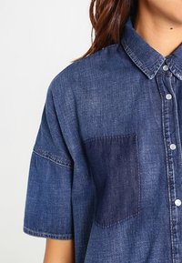 Denim-paita lyhyillä hihoilla, vaaleansinisiä ja tummansinisiä sävyjä, levitetty kaulus ja etutasku. Varustettu metallisilla napitusnapeilla.