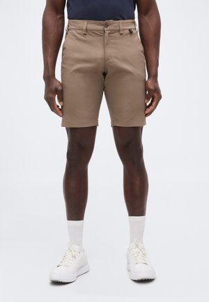 Homme portant un short beige longueur genoux, des chaussettes blanches et des baskets blanches, debout les bras détendus le long du corps sur un fond uni.