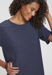 Junge Frau mit lockigem Haar, die ein marineblaues, geripptes Kurzarmshirt trägt, lächelt und mit einer Hand ihr Haar berührt.