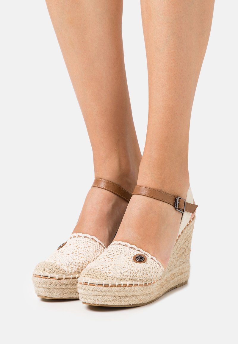 Espadrilles compensées avec empeigne en crochet beige, bride de cheville marron avec boucle, et semelle plateforme en jute portées pieds nus.
