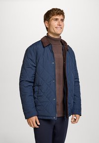 Marineblå quiltet jakke med brun fløjlsribskrave, knaplukning og sidelommer, båret over en struktureret brun sweater.