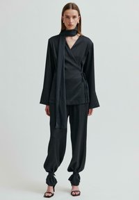 Černý top se zavazováním na krku, dlouhými rukávy a vázacím šátkem; doplněno širokými černými kalhotami, staženými u kotníků pomocí tkanic.