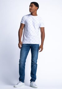 Tricou alb cu modele mici albastre, guler rotund, mâneci scurte, purtat cu blugi bleumarin și adidași albi. Stil casual, textură netedă.
