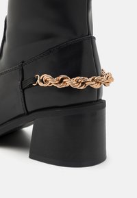 Bottine en cuir noir avec talon bloc et accent de chaîne tressée dorée sur le côté, mettant en valeur la texture et les détails du design.