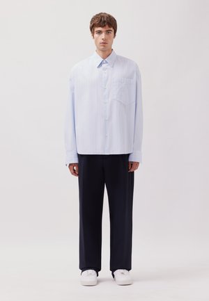 Camicia a righe blu chiaro con colletto a punta, maniche lunghe e taschino sul petto, abbinata a pantaloni blu navy larghi e sneakers bianche.