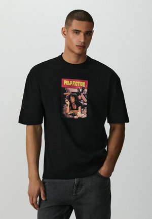 Mann trägt ein schwarzes T-Shirt mit einem Aufdruck des Filmplakats von "Pulp Fiction" und steht mit einer Hand in der Tasche vor einem einfarbigen hellen Hintergrund.