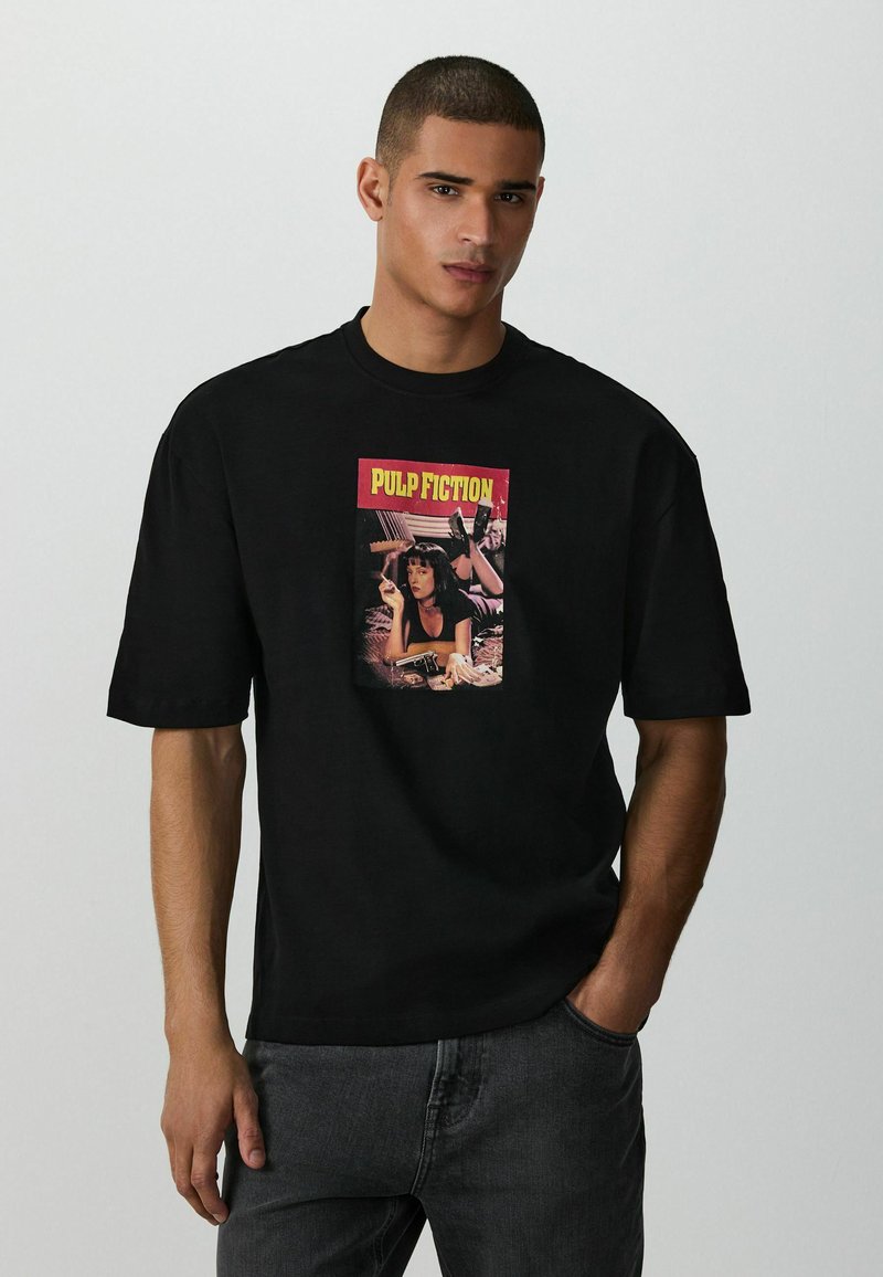 Homme portant un t-shirt noir avec une impression d'affiche du film "Pulp Fiction", debout une main dans la poche sur un fond clair uni.