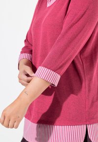 Fuchsia långärmad topp med V-ringning, med rosa och vita randiga manschetter och ett lagerutseende vid fållen. Mjuk, texturerad tyg.