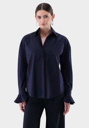 Camicia blu navy a maniche lunghe con bottoni, colletto a punta, polsini svasati e texture liscia. Il design è sartoriale e aderente.