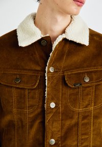 Veste en velours côtelé marron avec un col en shearling blanc, deux poches avant et des boutons en métal argenté avec le logo de la marque.