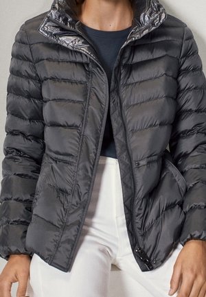 Personne portant une doudoune noire matelassée par-dessus un haut noir et un pantalon blanc, la veste partiellement dézippée avec les mains près des poches.