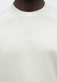 Primo piano di una persona che indossa una felpa grigio chiaro con la scritta "BOSS" ricamata in bianco sul petto.