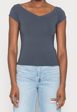 Vrouw draagt een nauwsluitende donkergrijze off-shoulder top en lichtblauwe gescheurde spijkerbroek, staand tegen een effen achtergrond.