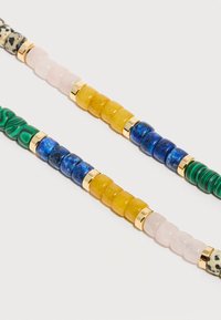 Deux bracelets de perles avec des pierres vertes, bleues, jaunes, roses, noires et blanches séparées par des perles intercalaires de couleur dorée sur fond blanc.