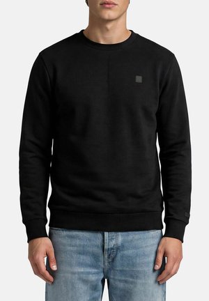 Mann trägt einen schlichten schwarzen Langarmpullover und hellblaue Jeans, steht vor weißem Hintergrund.