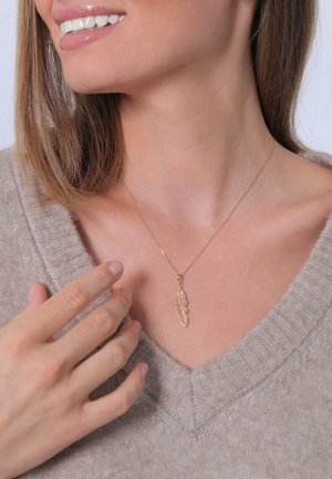 Goldfarbene Halskette mit einem Blätteranhänger, vor einem hellbeigen Pullover mit V-Ausschnitt. Einfaches Kettendesign, glatte Textur.