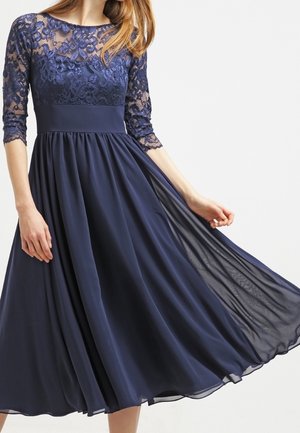 Laivastonsininen midimekko, jossa pitsinen yläosa, kolme neljänneksen hihaa, vartalonmyötäinen vyötärö ja ilmava sifonkihame, jonka naisen kevyesti nostama helma liehuu.