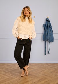 Světlé béžové žebrované pletené vesty přes bílou halenku s krajkovými detaily, kombinované s tmavými širokými džínami a růžovými podpatky. Modrá džínová bunda visí.