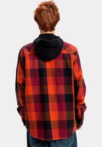 Camisa de franela a cuadros roja y negra con mangas largas, que presenta una sudadera negra debajo. Textura suave, diseño casual y cierres de botones.