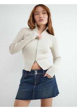 Kobieta w beżowym, prążkowanym swetrze na zamek, częściowo rozpiętym, oraz ciemnoniebieskiej dżinsowej mini spódnicy na jasnym jednolitym tle.