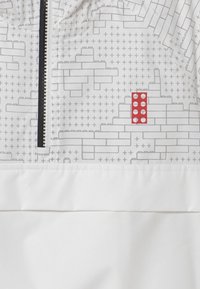 Vit tröja med grå tegelmönster och rutiga detaljer, med framdragkedja, huva och en distinkt röd rektangulär logotyp.