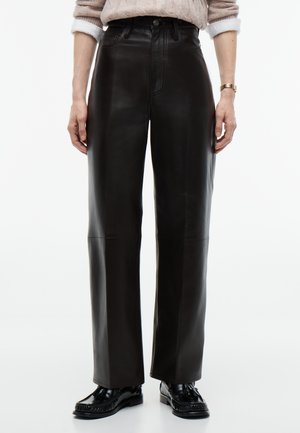 Personne portant un pantalon large en cuir noir taille haute, des mocassins noirs, un pull beige en maille torsadée et une montre-bracelet dorée.