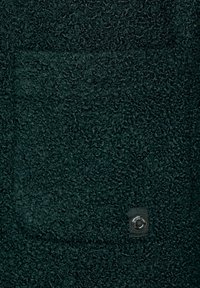 Tessuto verde scuro con una superficie morbida e pelosa, dotato di una piccola tasca a toppa e di un dettaglio in metallo di colore argento.