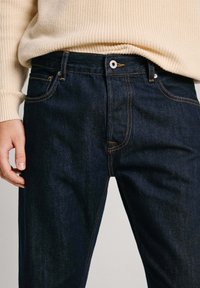 Jeans in denim blu scuro con cuciture arancioni, design a cinque tasche e chiusura con zip frontale. Il materiale presenta una leggera texture e brillantezza.