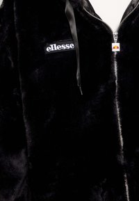 Sudadera con capucha de felpa negra con el logotipo blanco de "ellesse" en el pecho y tirador de cremallera de la marca, llevada sobre una prenda blanca.