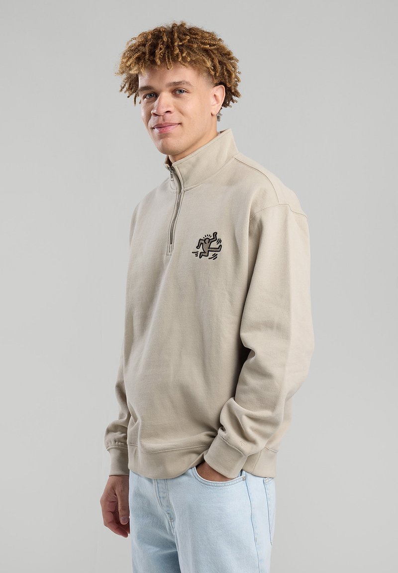 Beige fleece pullover with a high collar and half-zip. Features a black embroidered design on the left chest and ribbed cuffs.