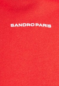 Tissu rouge avec le texte blanc brodé "SANDRO PARIS" centré sur le matériau.