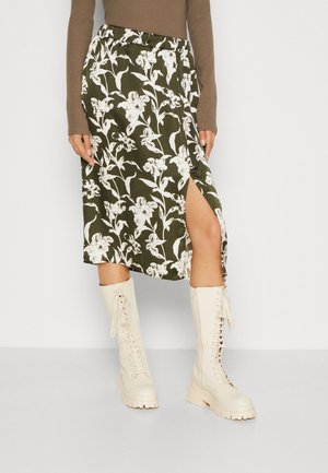 Kvinde iført olivengrøn blomsterprintet midi-nederdel med front-split, brun langærmet top og cremefarvede snørestøvler til knæene.