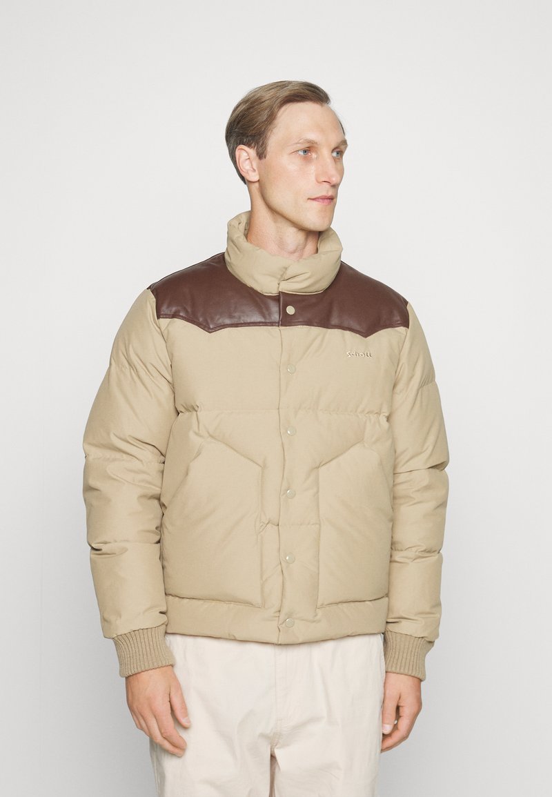Giacca puffer beige con dettagli in pelle marrone sulle spalle, chiusura a scatto, polsini a costine e texture trapuntata. Due tasche frontali.