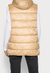 Beige pufferväst med kviltade sektioner och huva, tillverkad av glänsande syntetiskt material. Bärs över en ljusgrå långärmad tröja.
