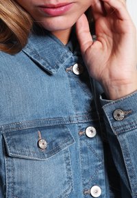 Chamarra de denim de color azul claro con herrajes plateados, que cuenta con dos bolsillos en el pecho y detalles de costura visibles alrededor del cuello y los botones.