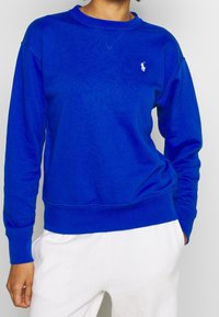 Blå bomulls sweatshirt med rundhals, ribbade ärmslut och nederdel, med en liten vit broderad logotyp på bröstet.