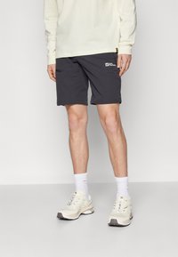 Man draagt zwarte Jack Wolfskin korte broek, wit shirt met lange mouwen, witte sokken en beige hardloopschoenen, staand tegen een effen achtergrond.