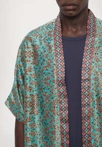 Kimono-lignende kimono i teal silke med indviklede sorte blomster mønstre. Har en rød og blå geometrisk kant langs de åbne kanter.