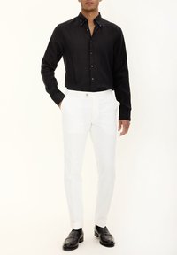 Schwarzes Langarmhemd mit Button-Down-Kragen, kombiniert mit weißen maßgeschneiderten Hosen und schwarzen Lederloafers, vor einem neutralen Hintergrund.