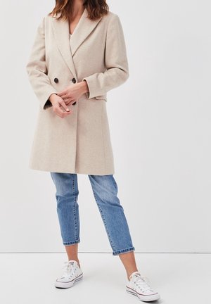 Femme portant un manteau beige à double boutonnage, un jean bleu raccourci et des baskets blanches basses, debout devant un fond clair uni.