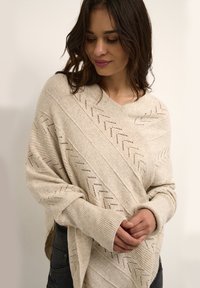 Beige knitted wrap with a V-neck, featuring openwork zigzag patterns and ribbed cuffs. Soft texture with a loose, oversized fit.