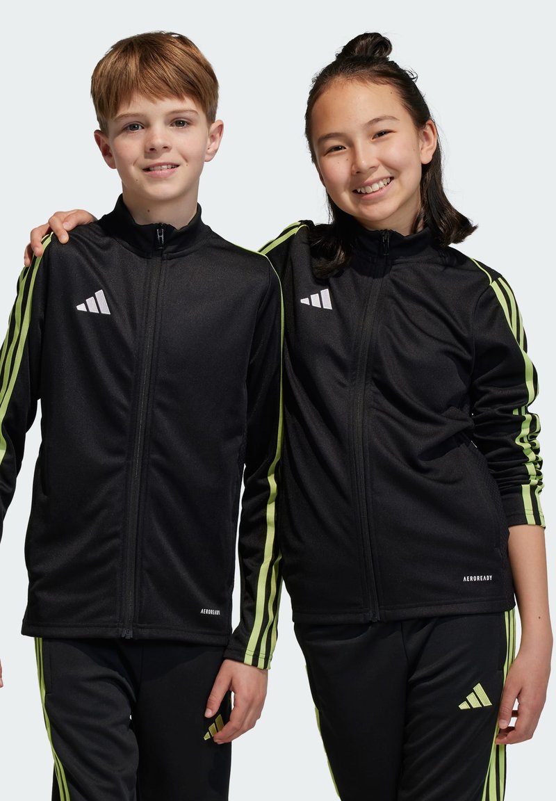 Zwarte sportjacks met groene strepen, logo-accenten op de borst, gecombineerd met bijpassende zwarte broeken. Beide jacks hebben een rits aan de voorkant.