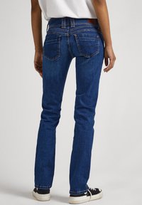 Jeans ajustados en denim azul oscuro con una cintura de altura media, cinco bolsillos y costuras anaranjadas. Combinados con zapatillas negras, vistos desde atrás.