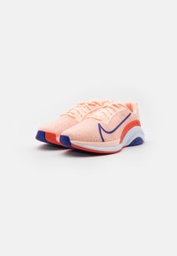 Un paio di sneaker sportive color pesca con accenti blu e rossi, parte superiore in rete e suole bianche imbottite, posizionate su uno sfondo bianco semplice.