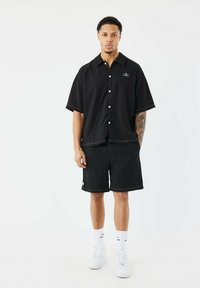 Camisa negra de manga corta con botones y textura vertical, combinada con pantalones cortos a juego. Ambos presentan costuras blancas y un pequeño detalle de logo.