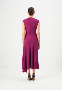 Donna con i capelli raccolti, che indossa un vestito viola senza maniche asimmetrico e sandali neri con tacco alto, in piedi davanti a uno sfondo neutro.