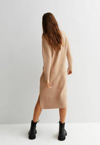 Robe beige en maille avec manches longues et ourlet côtelé, présentant une fente latérale. Associée à des bottes à plateforme noires.