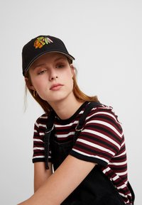 Black cap with embroidered head logo. Soft cotton striped shirt in red, white, and black. Overall made of dark denim with adjustable straps.