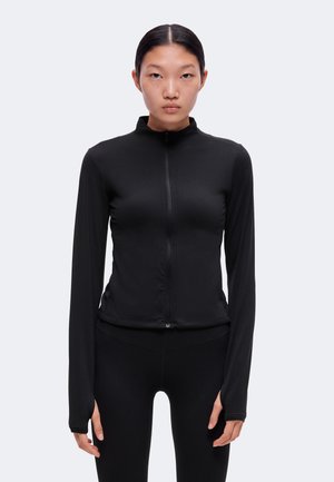 Mujer joven con el cabello oscuro recogido, lleva una chaqueta negra ajustada de manga larga con cremallera frontal y leggings negros a juego, mirando al frente.