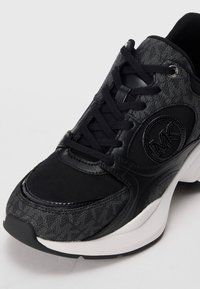 Black sneaker featuring a textured fabric upper, leather accents, a rounded toe, and a prominent logo on the side. White sole at the base.